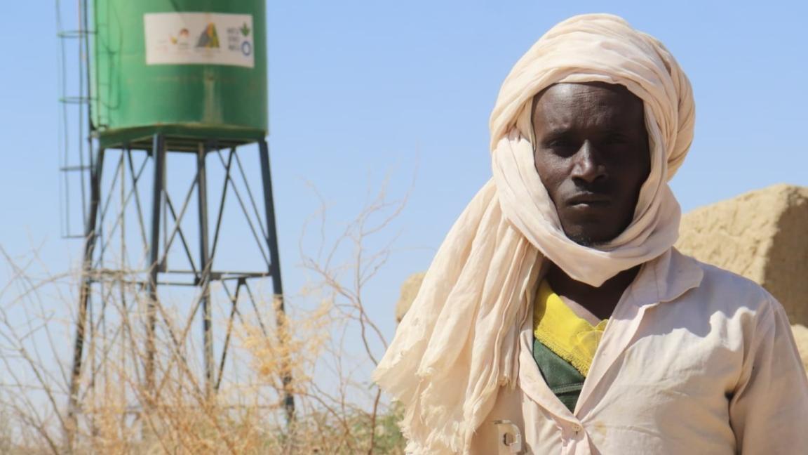 Ein Mann – Ibrahim Yatara – steht in Wüstenkleidung vor einem grünen Wassertank in einer trockenen Landschaft.