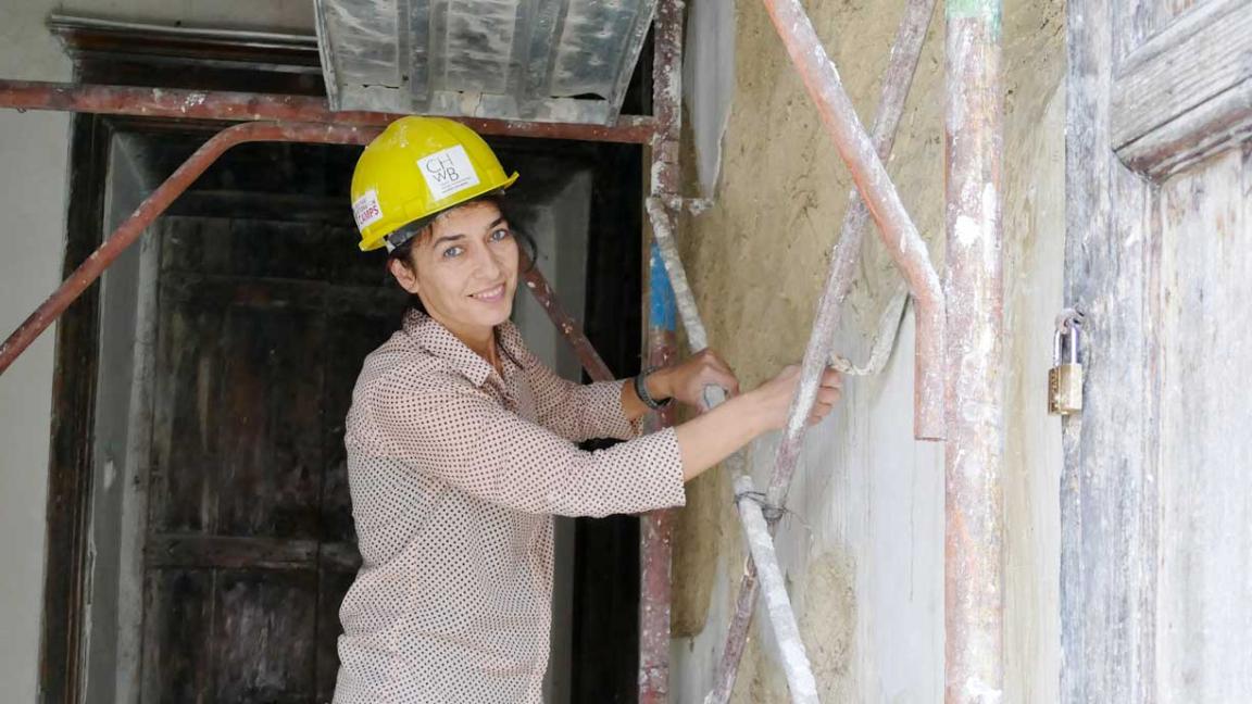 A woman aged around 30 with short dark hair and a yellow hard hat is plastering a wall in a room with exposed building fabric and an old door. She is wearing a polka dot shirt and smiles at the camera while standing on scaffolding.