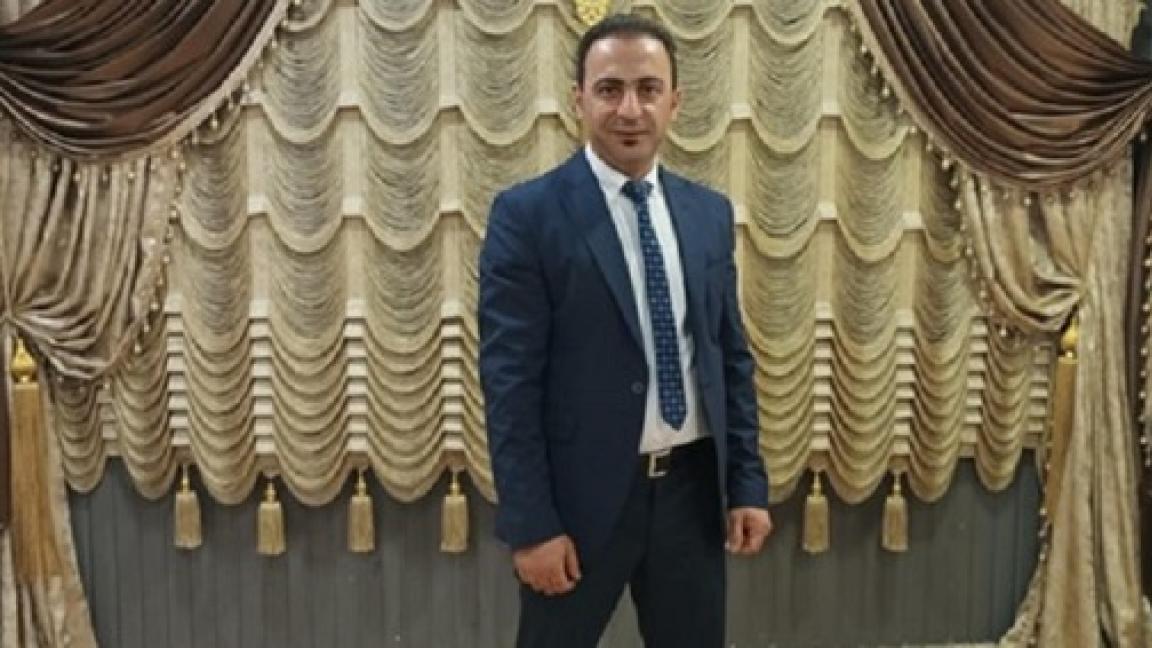 A man wearing a suit and tie is standing in front of artistically draped curtains in gold, beige and brown hues.