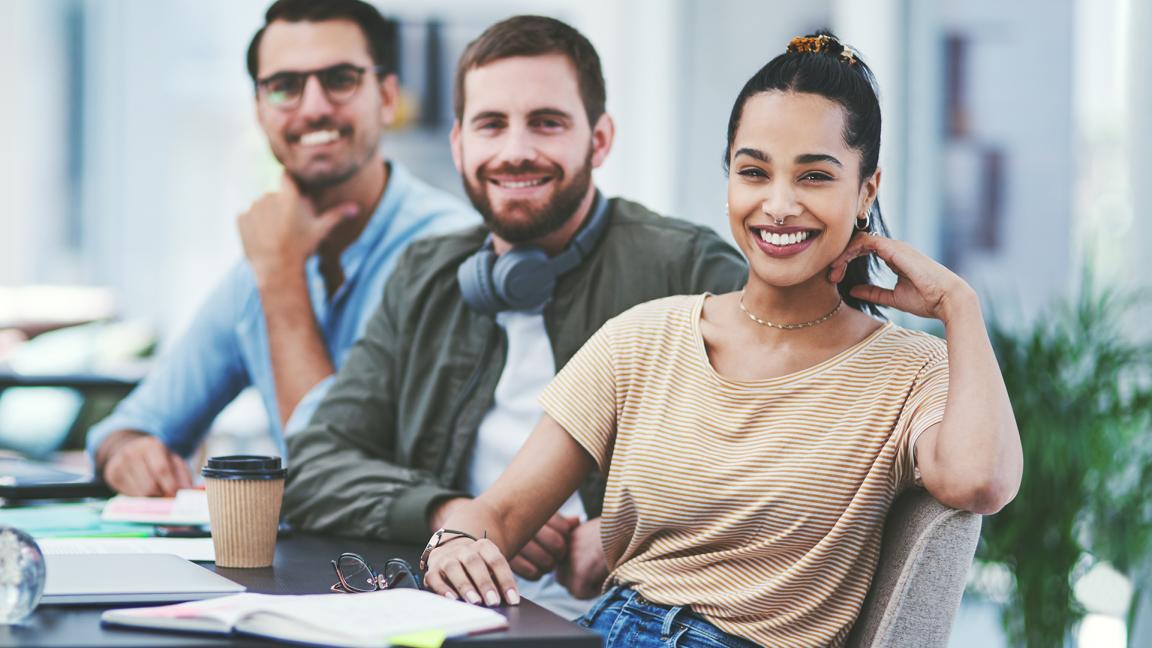 Drei junge Erwachsenen sitzen in einem Büro und schauen in die Kamera