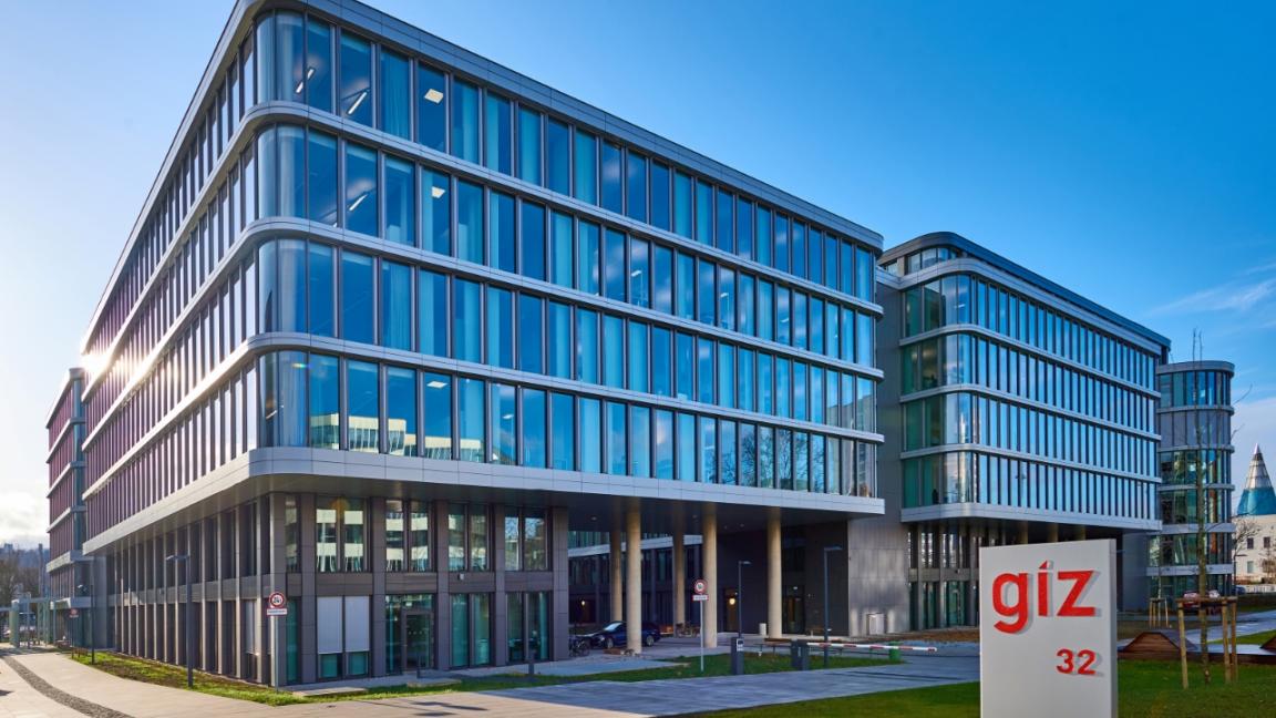 Gebäude mit vielen Fenstern vor einem sommerlich blauen Himmel. Im Vordergrund steht eine Stehle mit der Auschrift "giz" und "32"