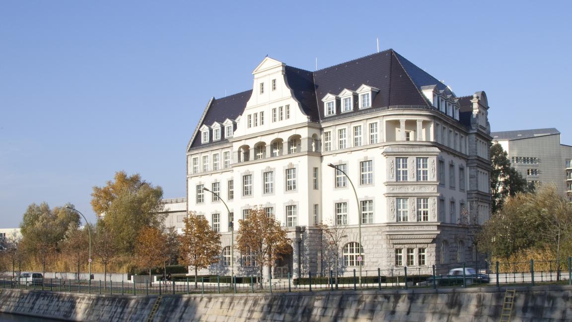 Historisches, mehrstöckiges Gebäude mit dunklem Schrägdach und zahlreichen Fenstern am Ufer eines Kanals; umgeben von Bäumen und modernen Gebäuden im Hintergrund, bei klarem Himmel.
