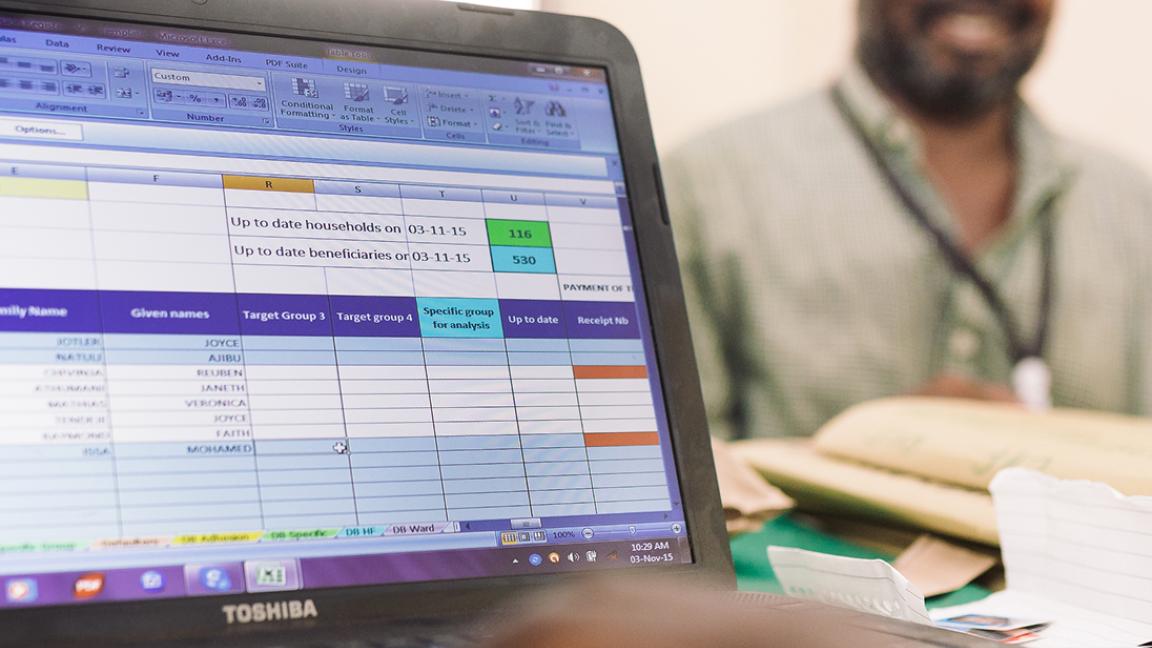 Ein Mensch tippt auf einem Laptop mit Excel-Tabelle, im Hintergrund sitzt ein lächelnder Mann an einem Schreibtisch.