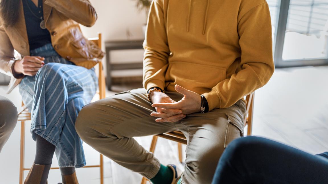 Teilweise sichtbare Menschen sitzen in einem Stuhlkreis in einer hellen Atmosphäre. Mit den Händen wird gestikuliert.