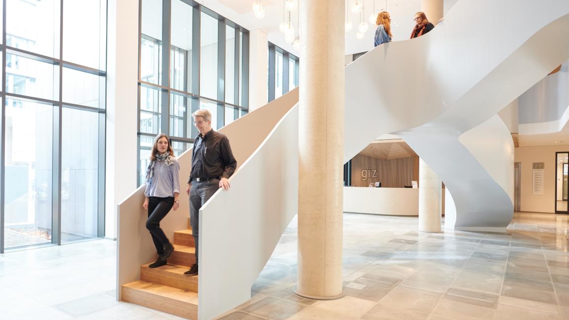 Heller und hoher Innenraum eines modernen Gebäudes mit einer Säule und einem organischen Treppenaufgang. Auf der Treppe laufen vier Personen.