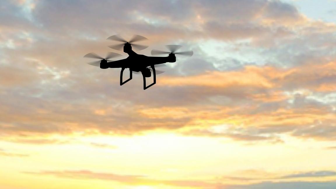 Die Silhouette einer Person, die eine schwebende Drohne per Fernsteuerung steuert, vor einem Abendhimmel