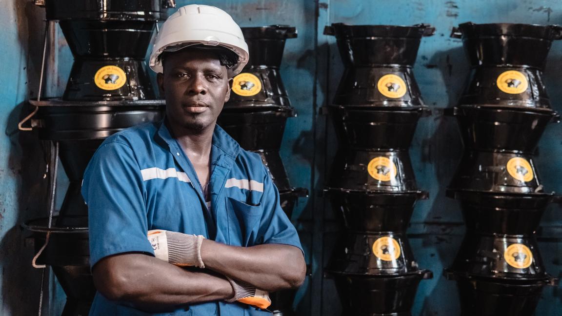 A man in a white helmet stands in front of a wall where cookers are piled up.