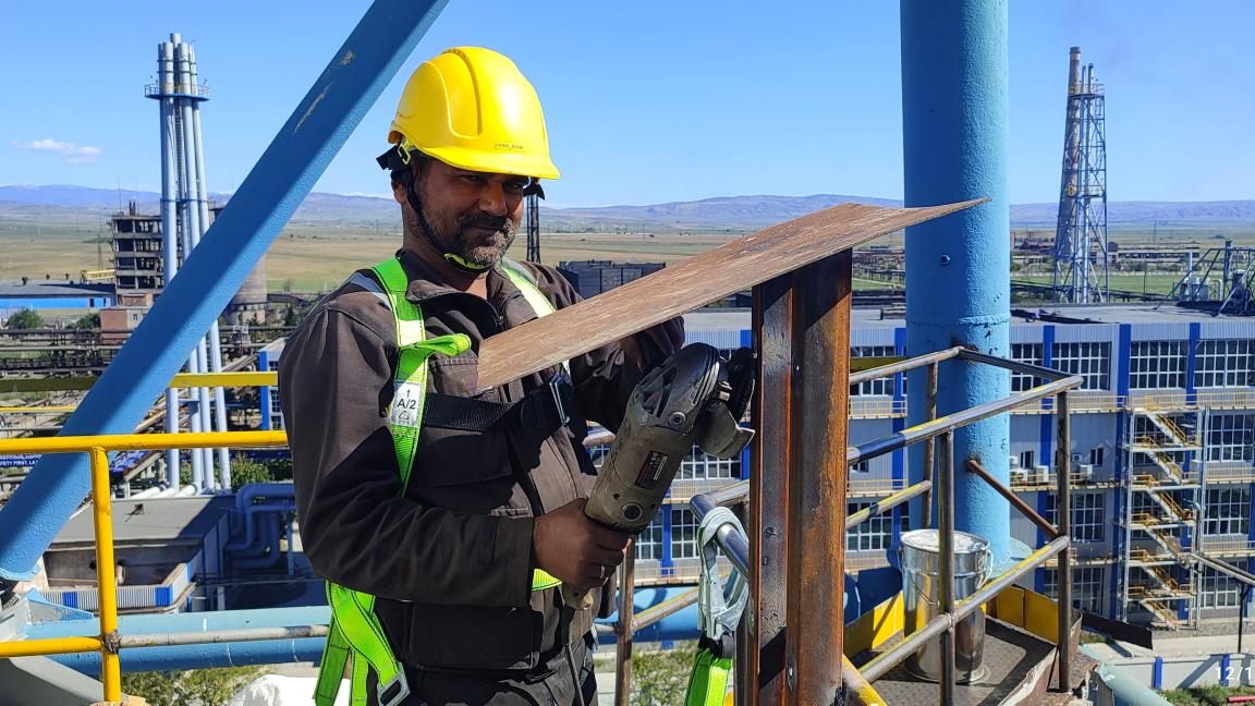 A man in a yellow helmet stands on a scaffold and grinds down a steel frame.
