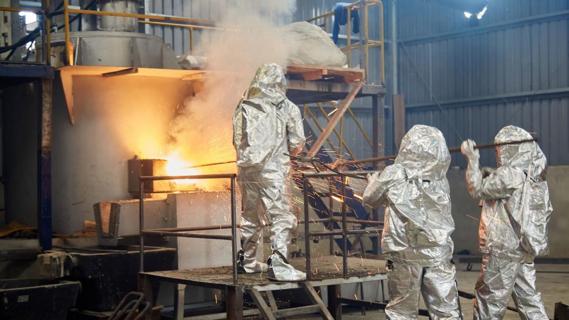 Drei Personen in silbernen Schutzanzügen arbeiten an einem glühenden Ofen in einer Zinnverarbeitungsanlage