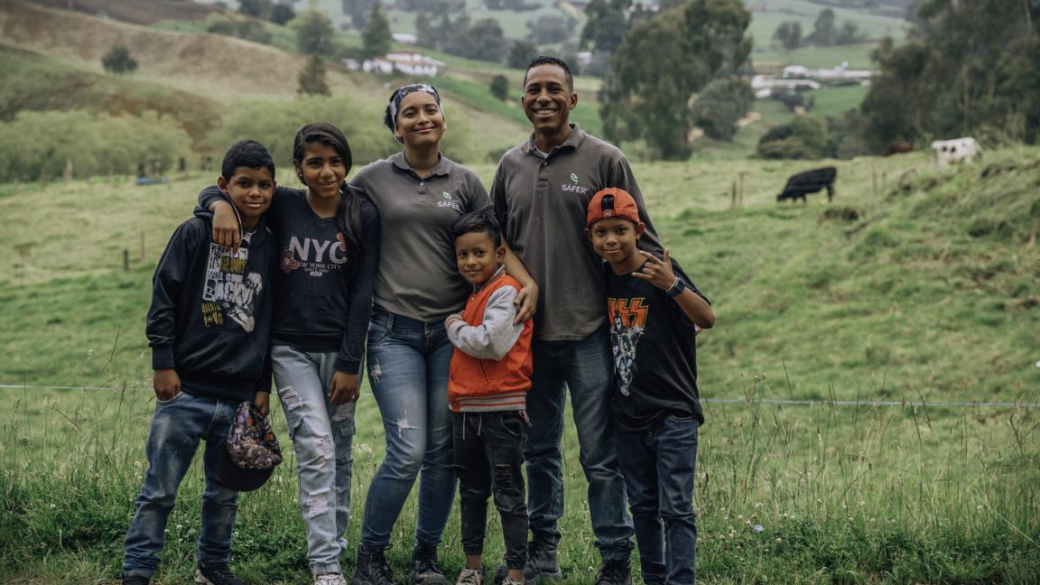Zwei Erwachsene und vier Kinder stehen Arm in Arm in einer Landschaft mit grünen Hügeln