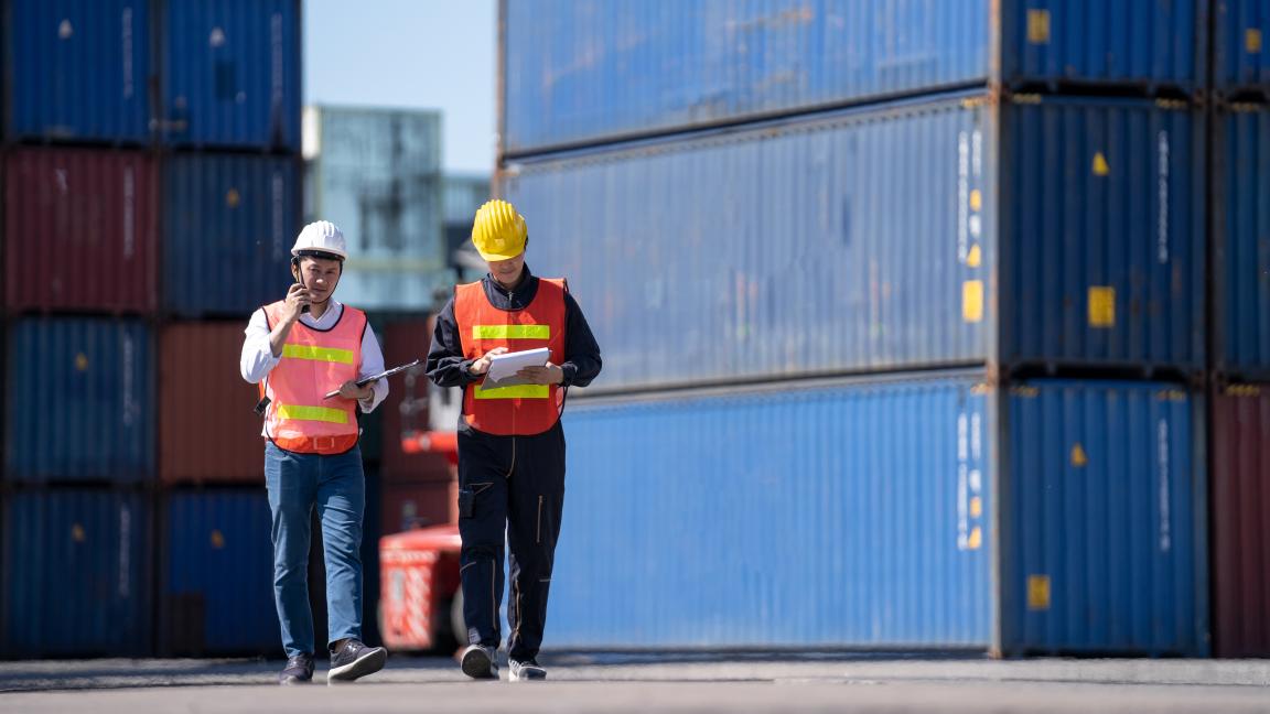 Zwei Männer laufen ein Hafengelände entlang.
