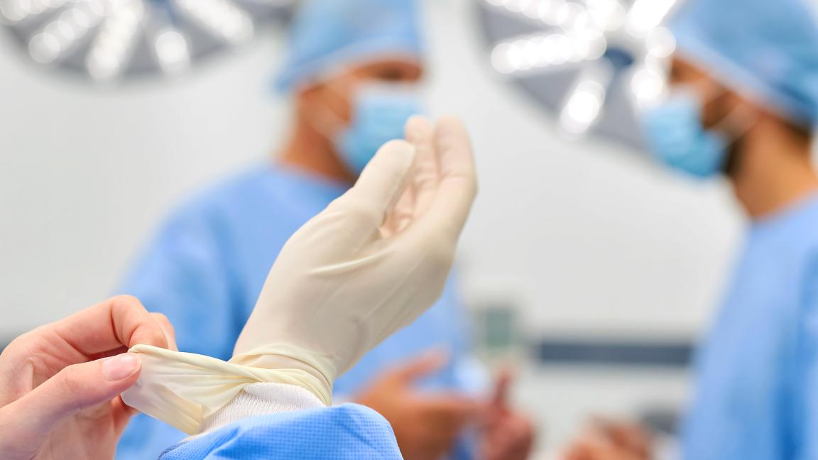 Eine medizinische Fachkraft zieht sich in einem Operationssaal sterile Handschuhe über, im Hintergrund sind unscharf zwei weitere Personen in OP-Kleidung zu sehen.
