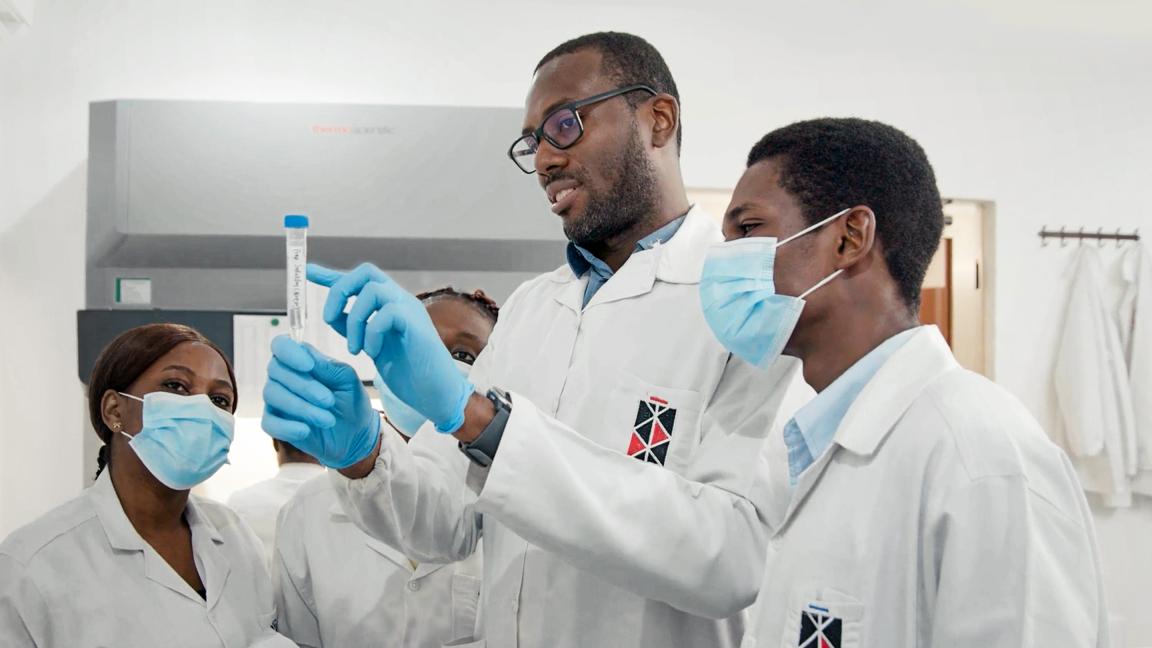 Ein Mann im Laborkittel zeigt einer Gruppe von Kollegen, wie man ein Testrohr mit einer Probe handhabt. Es wird Zusammenarbeit und Wissensaustausch im Labor betont.