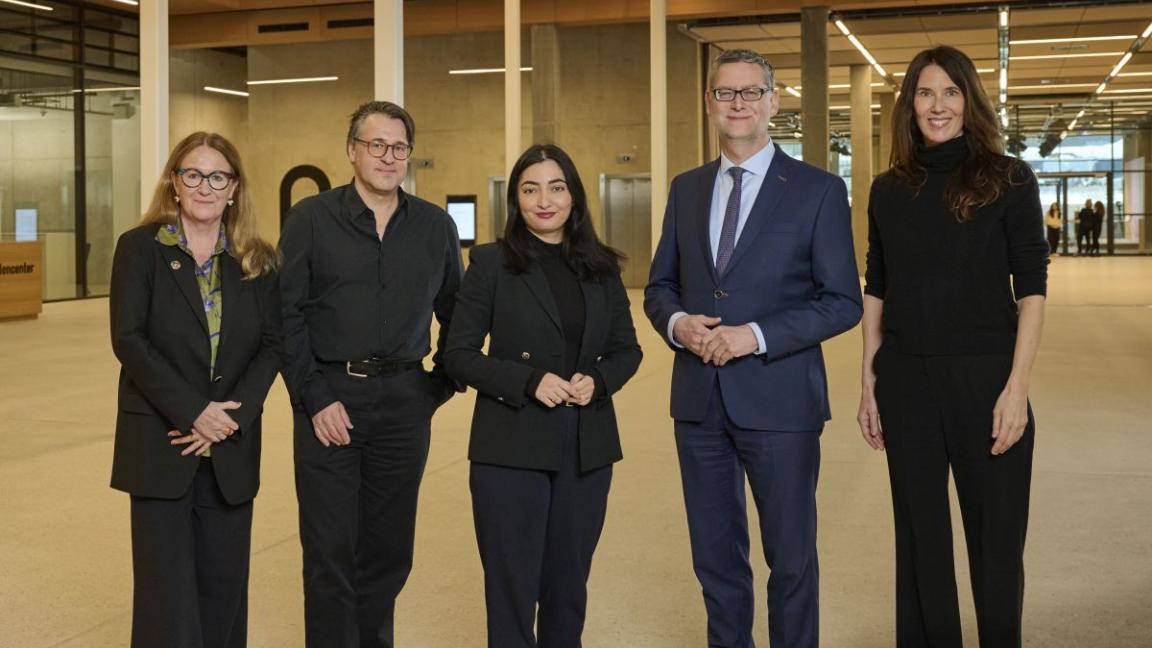 Five people in business attire are standing smiling in a large room.
