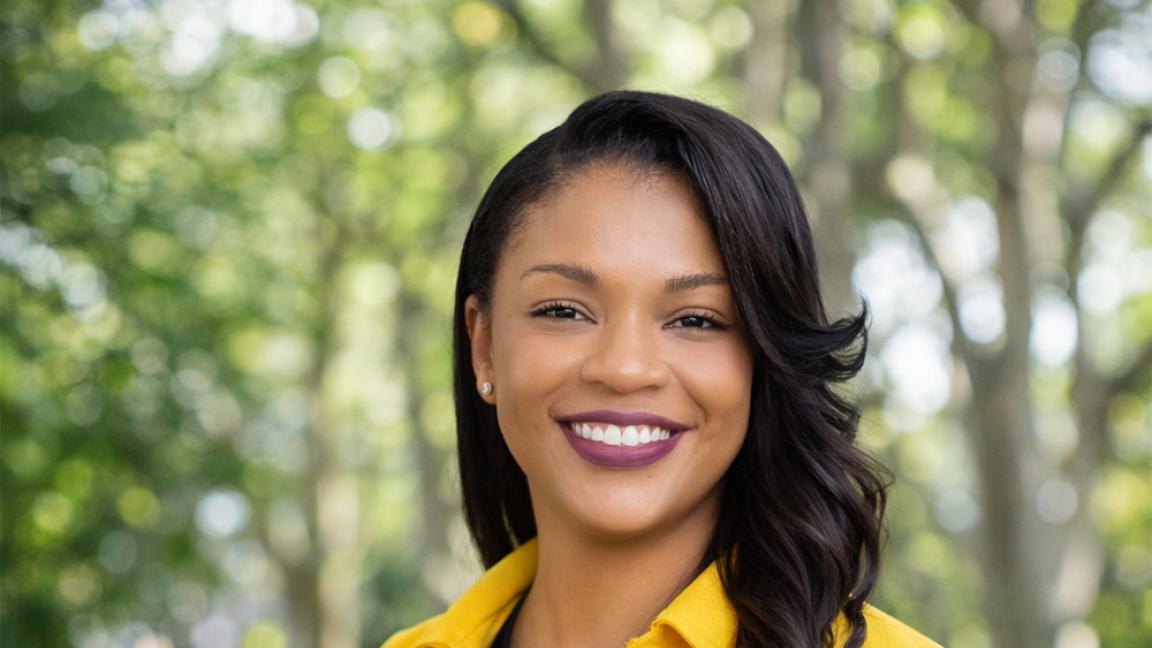 Porträt einer lächelnden Frau mit langen dunklen Haaren, gelbem Blazer und schwarzem Oberteil vor unscharfem grünen Parkhintergrund bei Tageslicht.