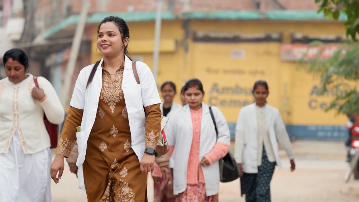 Eine Gruppe von Frauen in weißen Kitteln geht gemeinsam über eine Straße, angeführt von einer Frau in brauner Kleidung, vor einem unscharfen Gebäude im Hintergrund.