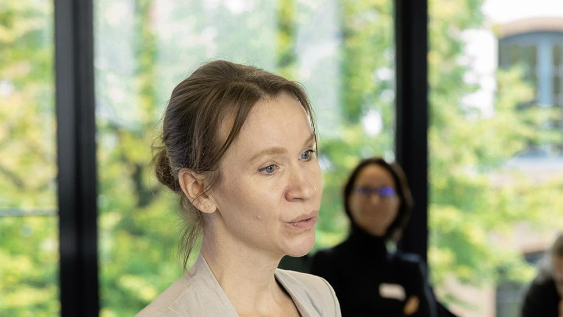 Frau mit zusammengebundenen Haaren hält ein Dokument in der Hand und spricht in einem hellen Seminarraum mit großen Fenstern, während im Hintergrund weitere Personen sitzen.