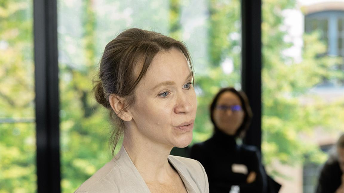 Woman with tied-back hair holding a document and speaking in a bright seminar room with large windows while other people are seated in the background.