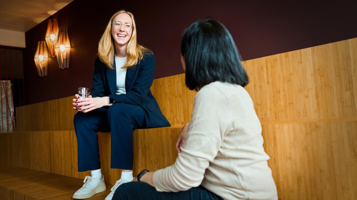 Zwei Personen sitzen in einem Innenraum auf Holzstufen und sprechen miteinander; eine Person hält ein Glas.
