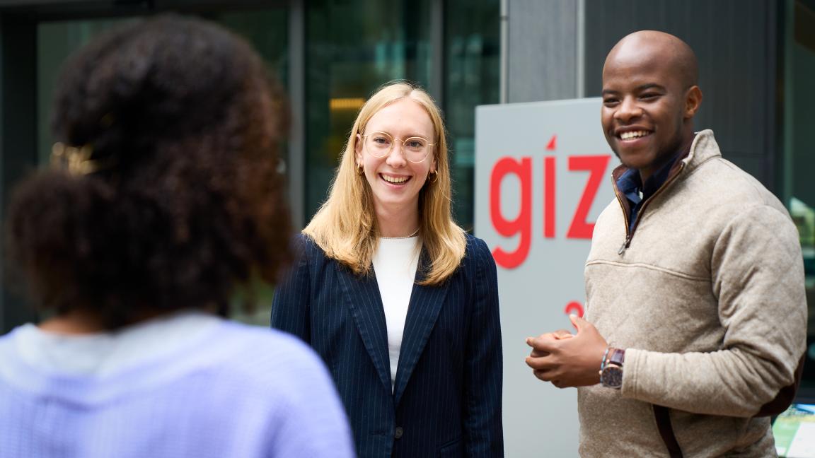 Zwei Personen stehen im Freien und sprechen; im Hintergrund ist ein Schild mit der Aufschrift „giz“ zu sehen.