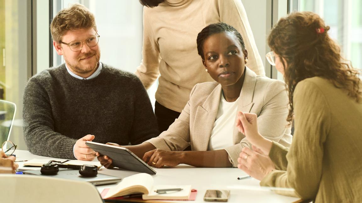 Several people sit at a table and discuss; a tablet and note-taking materials are on the table.