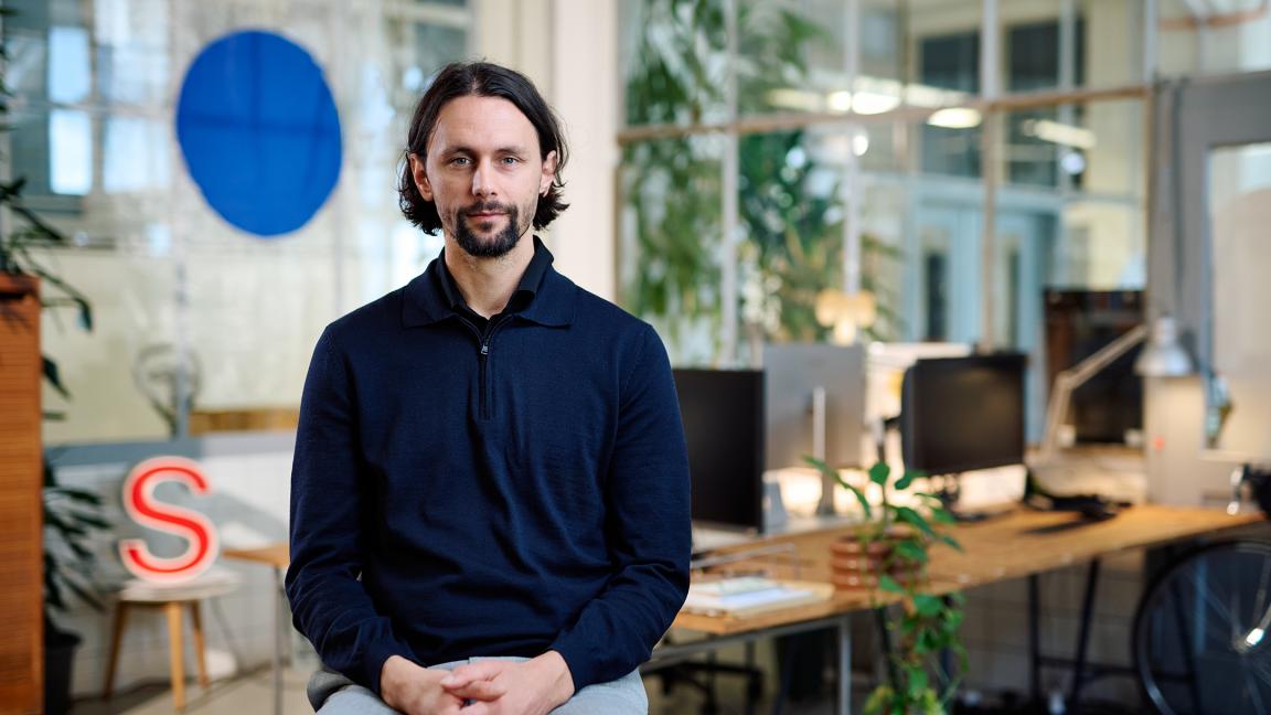 Ein Mann in einem dunkelblauen Pullover mit Reißverschluss sitzt mit gefalteten Händen in einem modernen, begrünten Büro und blickt ruhig in die Kamera.