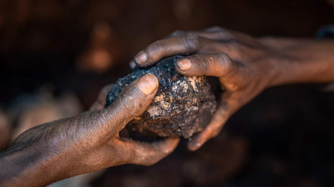 Zwei Hände halten einen dunklen, unbearbeiteten Gesteinsbrocken in einer schwach beleuchteten Umgebung.
