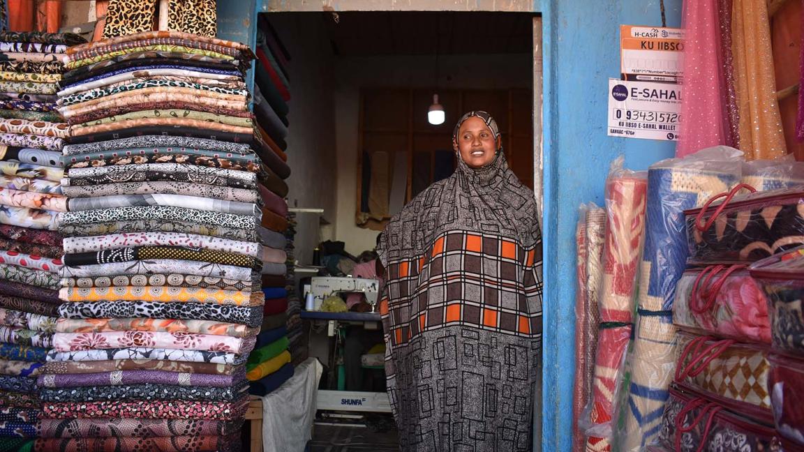 A woman in a colourful dress is standing next to a pile of fabrics in a small shop.