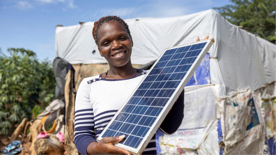 Eine Frau lächelt in die Kamera und hält vor einem provisorischen Unterstand ein Solarpanel in den Händen.