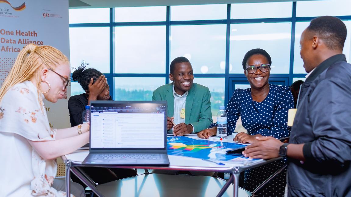 Mehrere Personen sitzen in einem hellen Raum an einem Tisch mit Laptop und Unterlagen und sprechen miteinander, im Hintergrund ein Banner „One Health Data Alliance Africa“.
