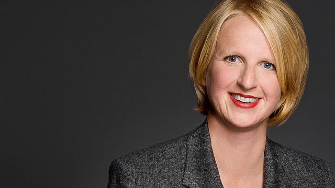 Porträt einer lächelnden Frau mit blondem Bob und rotem Lippenstift in grauem Blazer vor dunklem Hintergrund.