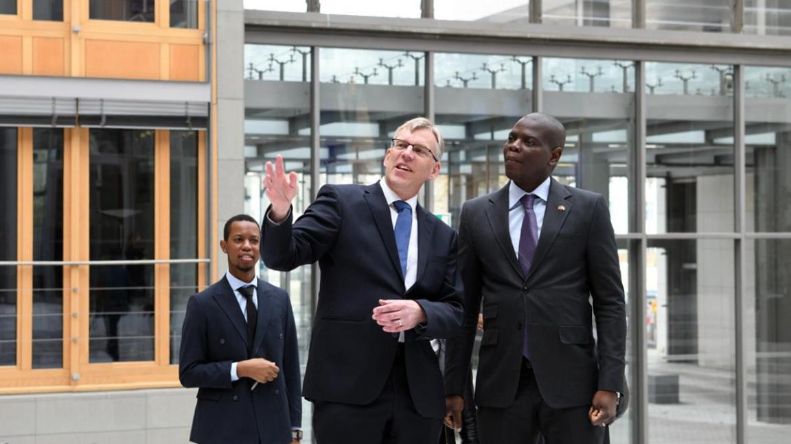 Drei Männer in Anzügen stehen in einem modernen Gebäude, während der Mann in der Mitte mit erhobener Hand gestikulierend nach oben zeigt.