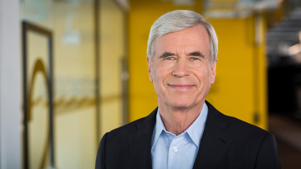 Portrait of an older gray-haired man in a suit in front of a yellow background.