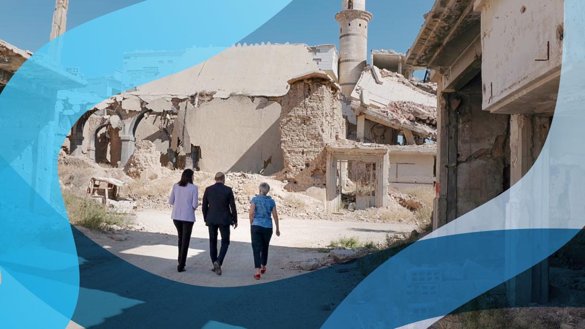 Three people walk along a street. All of the buildings are severely damaged. 