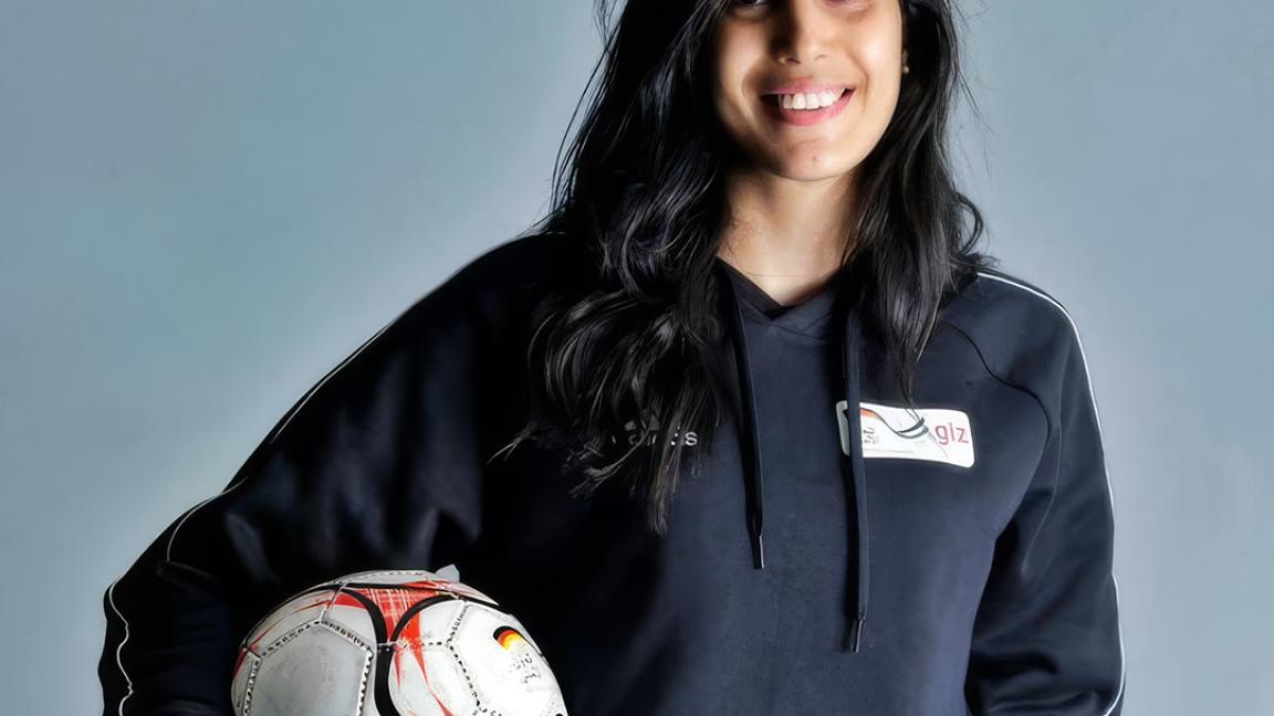Porträt einer lächelnden Person mit dunklen langen Haaren, die vor grauem Hintergrund einen Fußball unter dem Arm hält.
