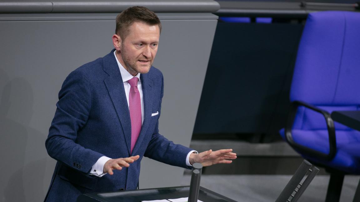 Ein Mann im blauen Anzug mit pinker Krawatte spricht gestikulierend an einem Rednerpult in einem Plenarsaal.