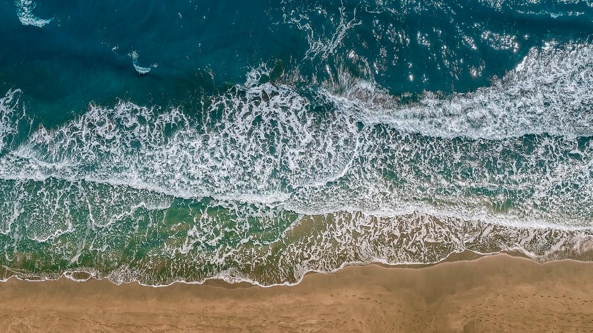 Aerial view of ocean waves breaking with white foam onto a golden-brown sandy beach, with turquoise to dark blue water in the background.