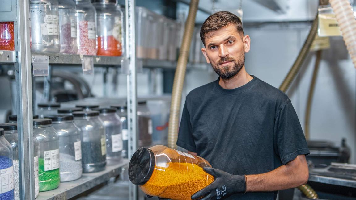 Ein bärtiger Mann in dunklem T-Shirt und schwarzen Arbeitshandschuhen hält ein Glas mit orangefarbenen Kunststoffgranulaten und blickt lächelnd in die Kamera, im Hintergrund stehen Regale mit weiteren Behältern voller farbiger Granulate.