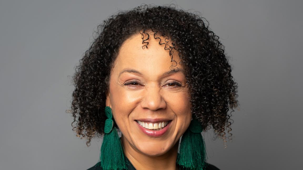 Portrait of a smiling woman with curly hair, a green top, a black blazer, and green tassel earrings against a neutral gray background.