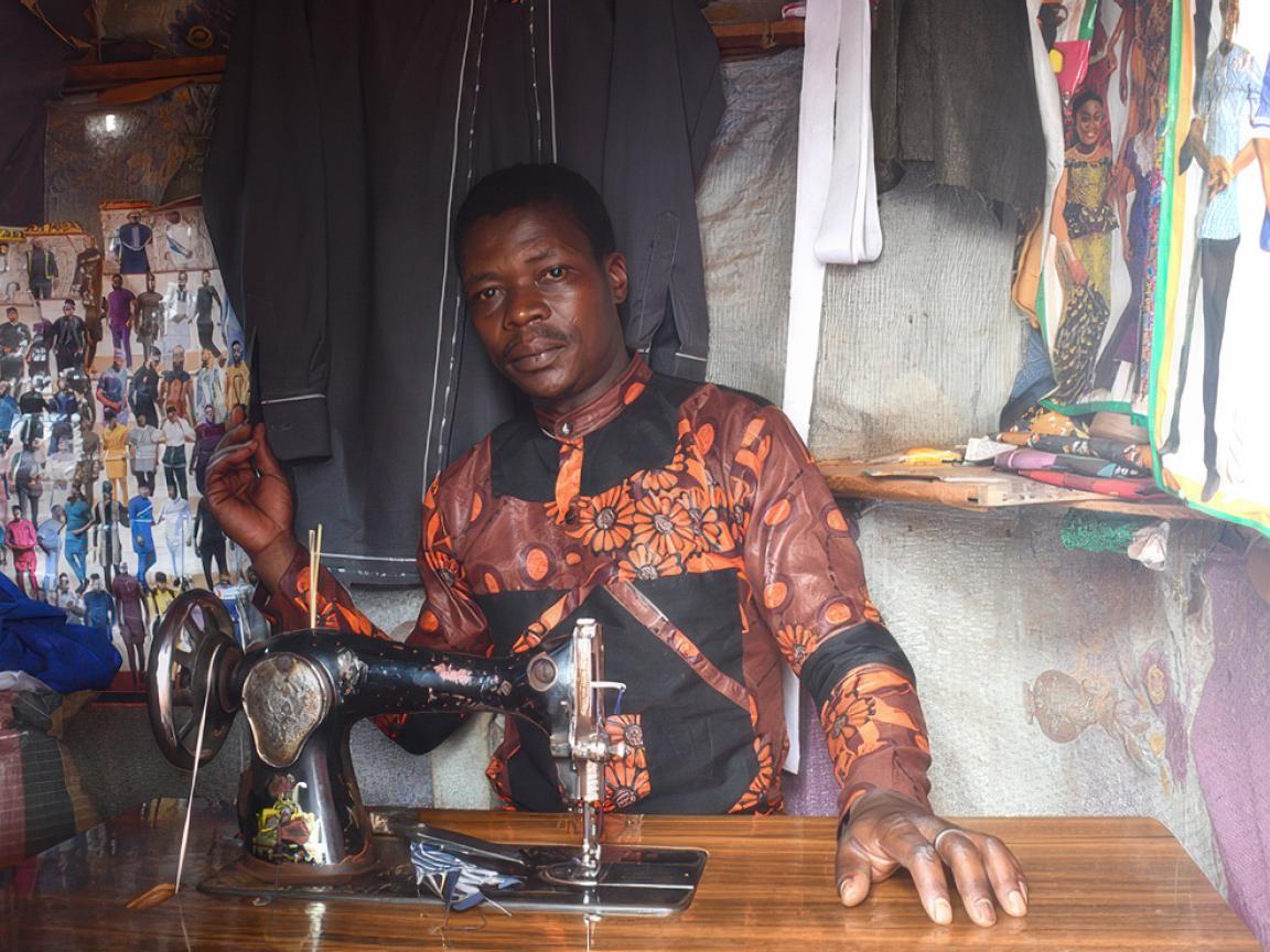 Ein Mann in einem kunstvollen Oberteil steht hinter einer Nähmaschine.