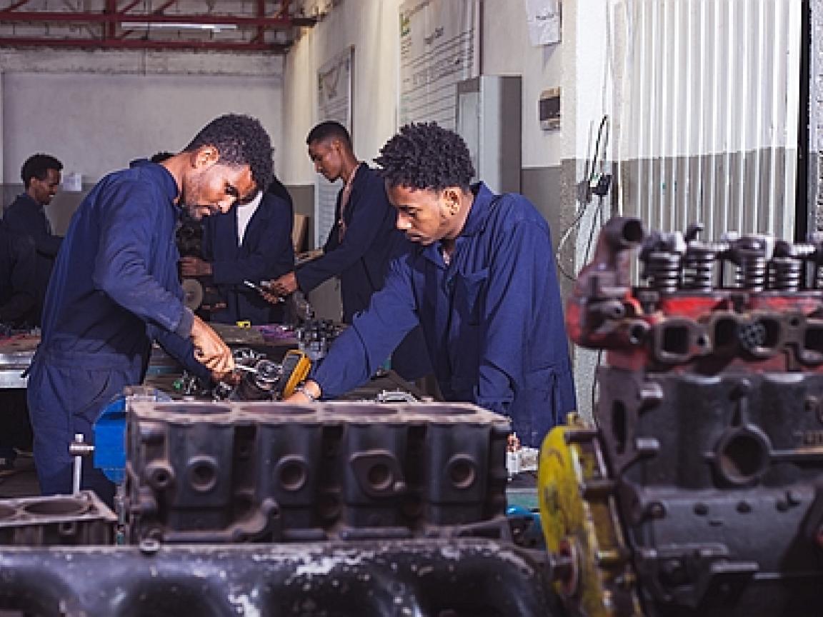 Two mechanics are working in a workshop. More mechanics are seen in the back.