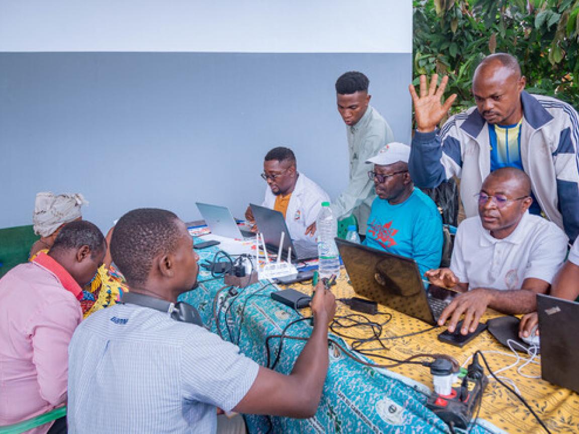 Vier Personen sitzen mit jeweils einen Laptop am Tisch. Gegenüber und dahinter stehen noch mehr Personen.