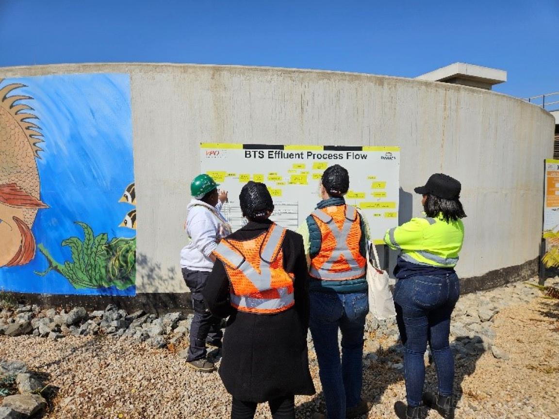 The picture shows four people from the back looking at a post on a cement wall. The poster has the headline "BTS Effluent Process Flow".