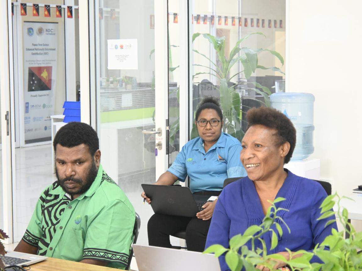 Three people are sitting in an office with laptops infront of them.
