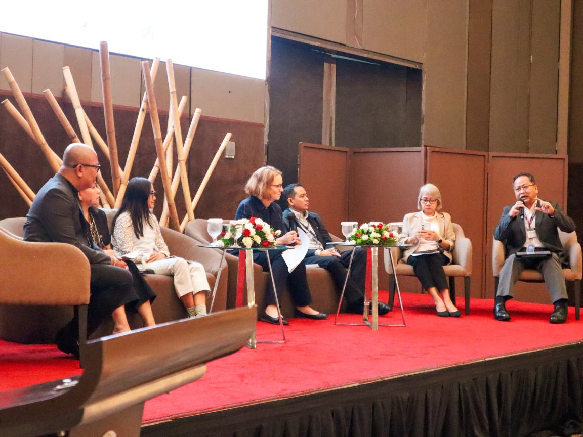 Six people are sitting at a podium discussion on a stage in chairs.