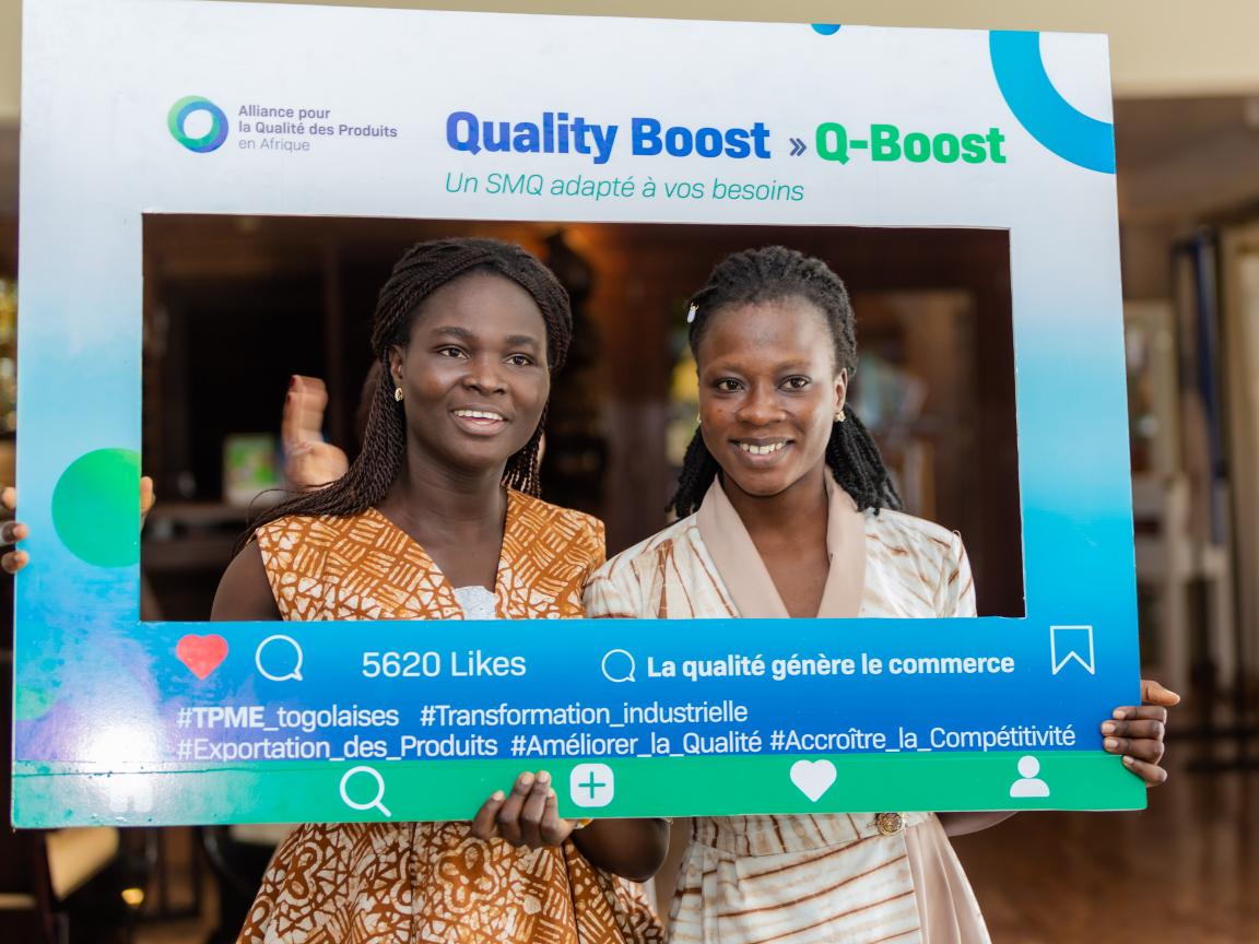 Two women smile while posing with a “Quality Boost (Q-Boost)” promotional frame.