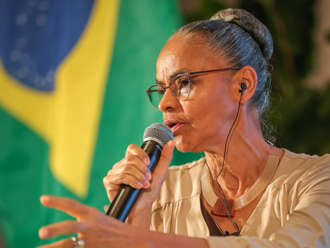 Eine Frau mit Mikrofon hält eine Rede vor einer Brasilienflagge.