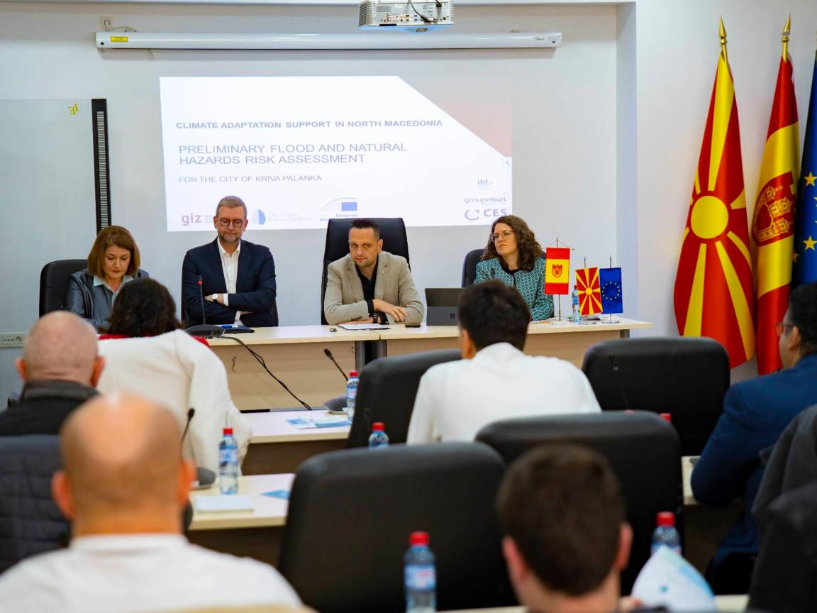 Vier Menschen sitzen bei einer Konferenz, rechts im Bild die Flagge Nordmazedoniens und der EU.