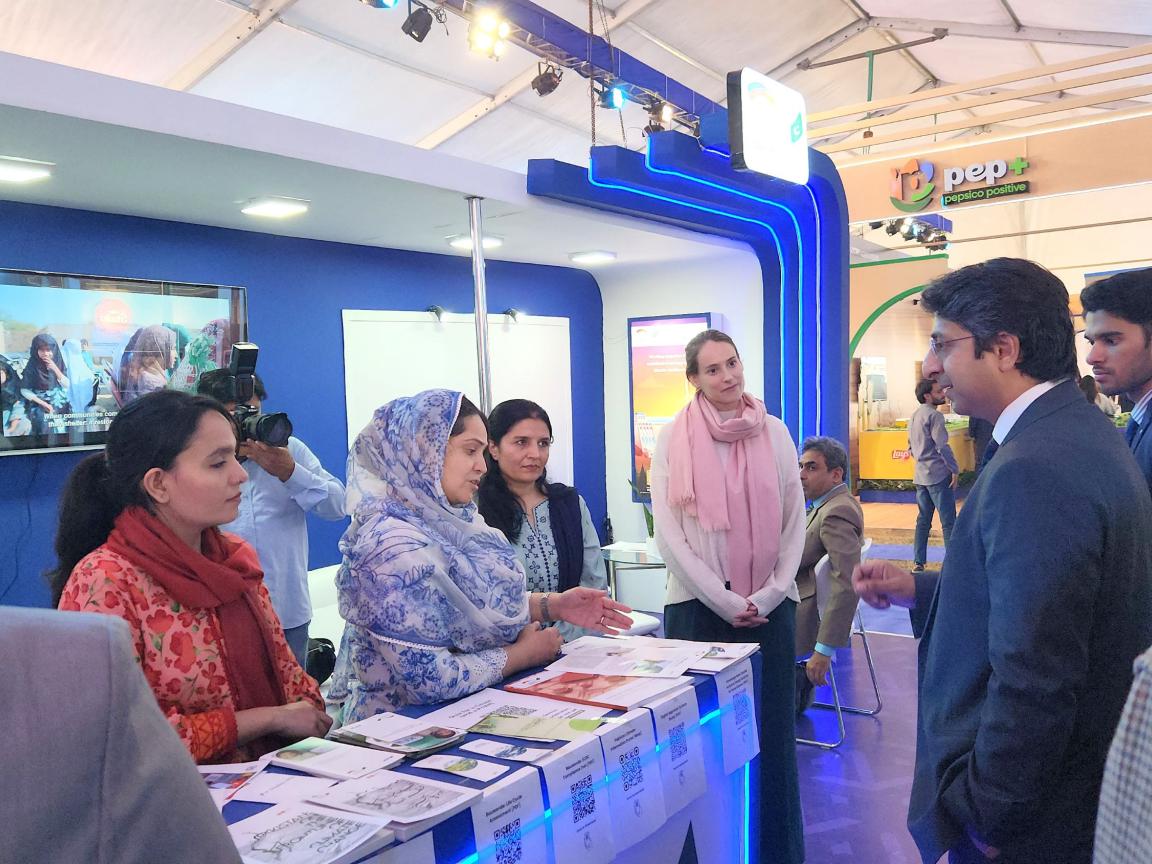 A group, including the PM's Coordinator, interacts with PGCEP staff at an exhibition booth featuring a display unit and brochures.