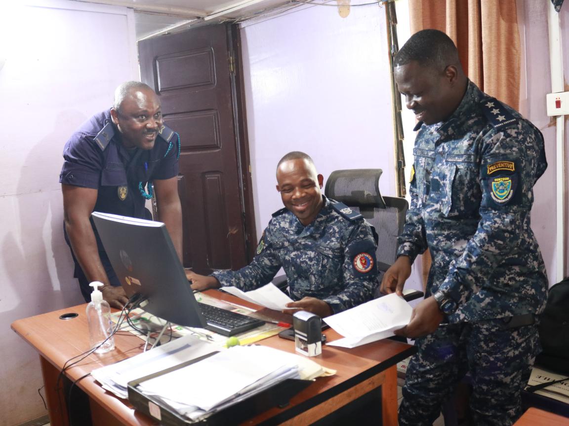 Three men in uniform are sitting at and standing around a desk.